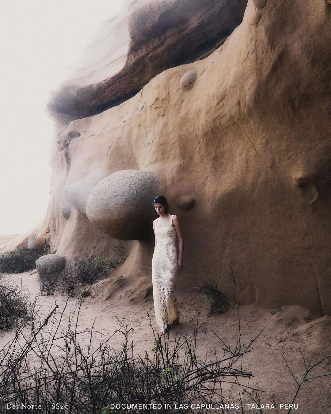 Adult female wearing long, light-colored dress stands on sand in arid landscape next to tan rock formation in editorial fashion shot.