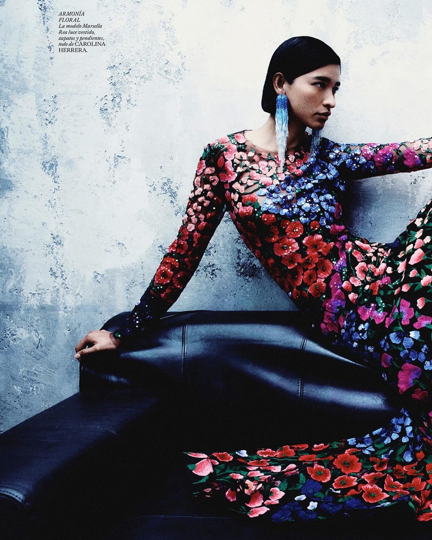 Fashion editorial shot: a woman poses on a black leather couch wearing a long-sleeved floral dress with blue earrings in front of a mottled wall.