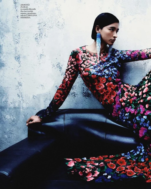 Fashion editorial shot: a woman poses on a black leather couch wearing a long-sleeved floral dress with blue earrings in front of a mottled wall.