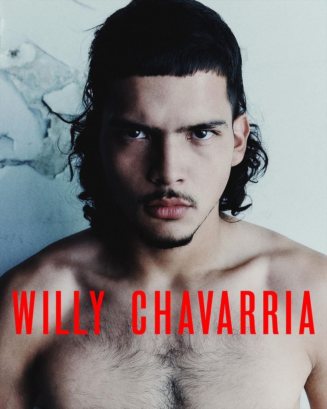 Portrait of a shirtless adult male with dark hair and a serious expression, against a textured off-white background and red text overlay.