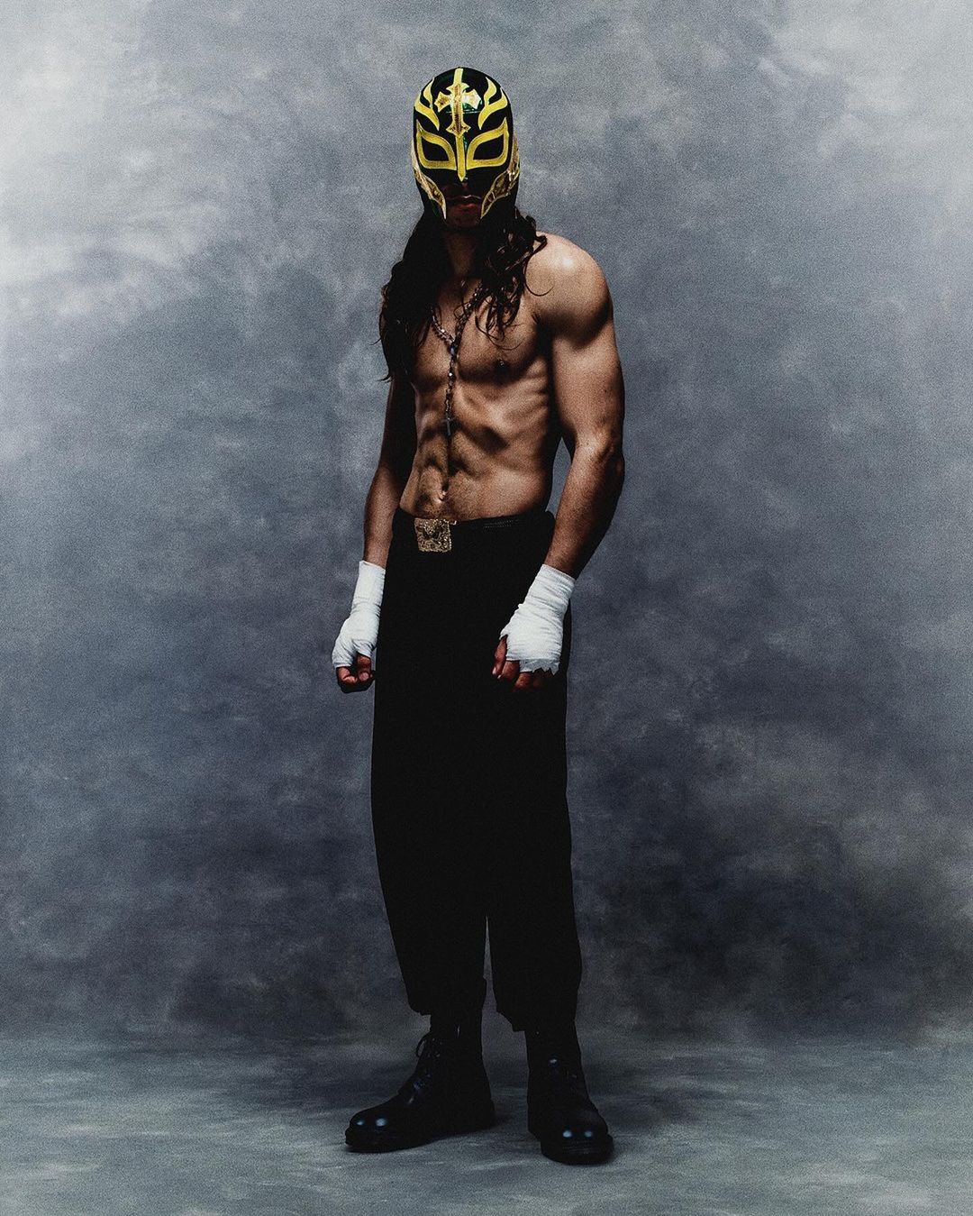 Muscular man wearing a lucha libre mask, hand wraps, and black pants poses in a full shot against a mottled grey backdrop.