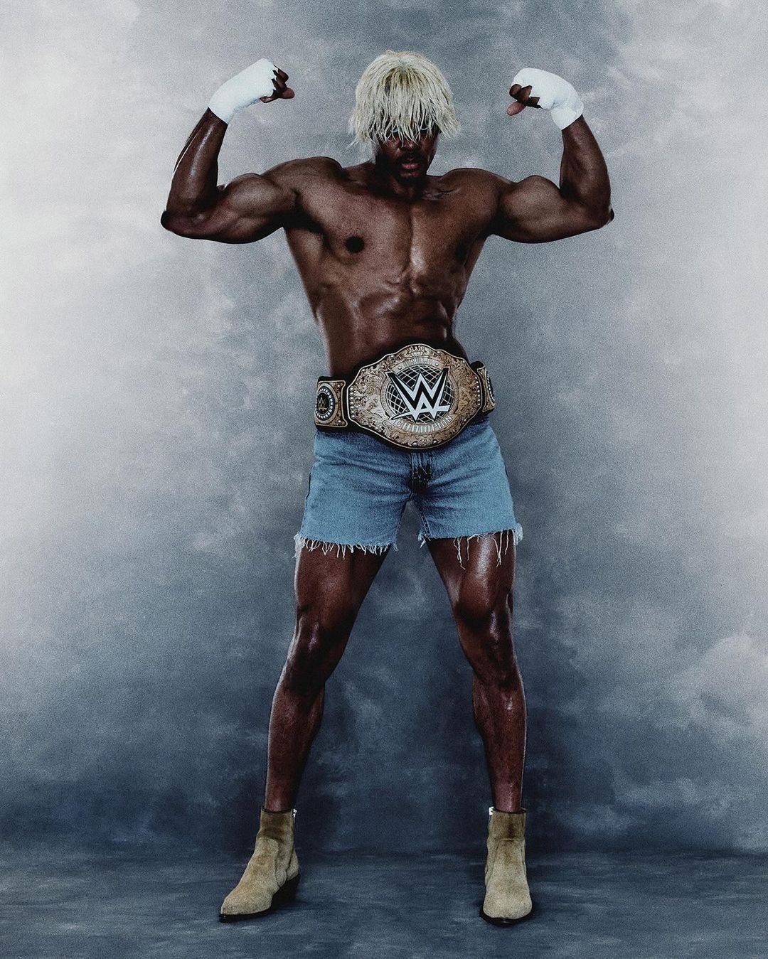 A muscular man with platinum hair poses in denim shorts and boots wearing a championship belt. He stands against a gray background.