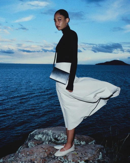 An adult female fashion model poses on a rocky outcrop at the ocean's edge wearing a black turtleneck, white skirt and ballet flats.