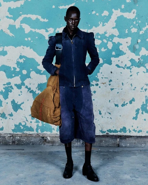 Dark-skinned model stands in a blue jacket, shorts, bag, socks and leather shoes against a distressed blue painted wall.