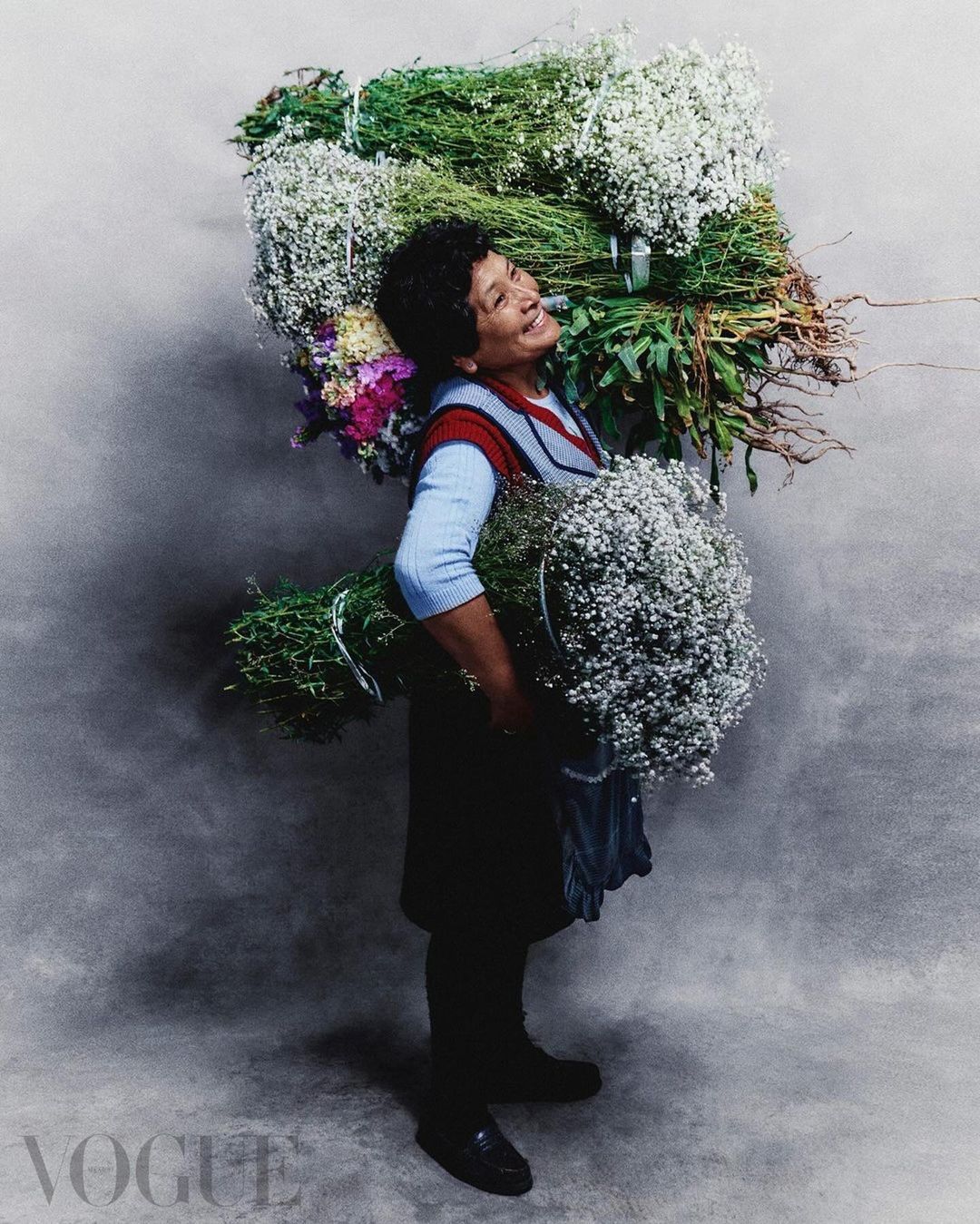 Portrait of a woman carrying and surrounded by large bundles of flowers against a grey background; fashion editorial for Vogue.