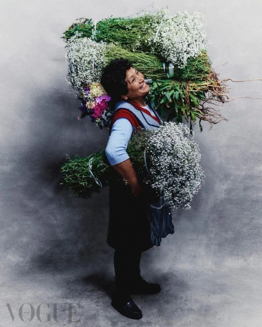 Portrait of a woman carrying and surrounded by large bundles of flowers against a grey background; fashion editorial for Vogue.
