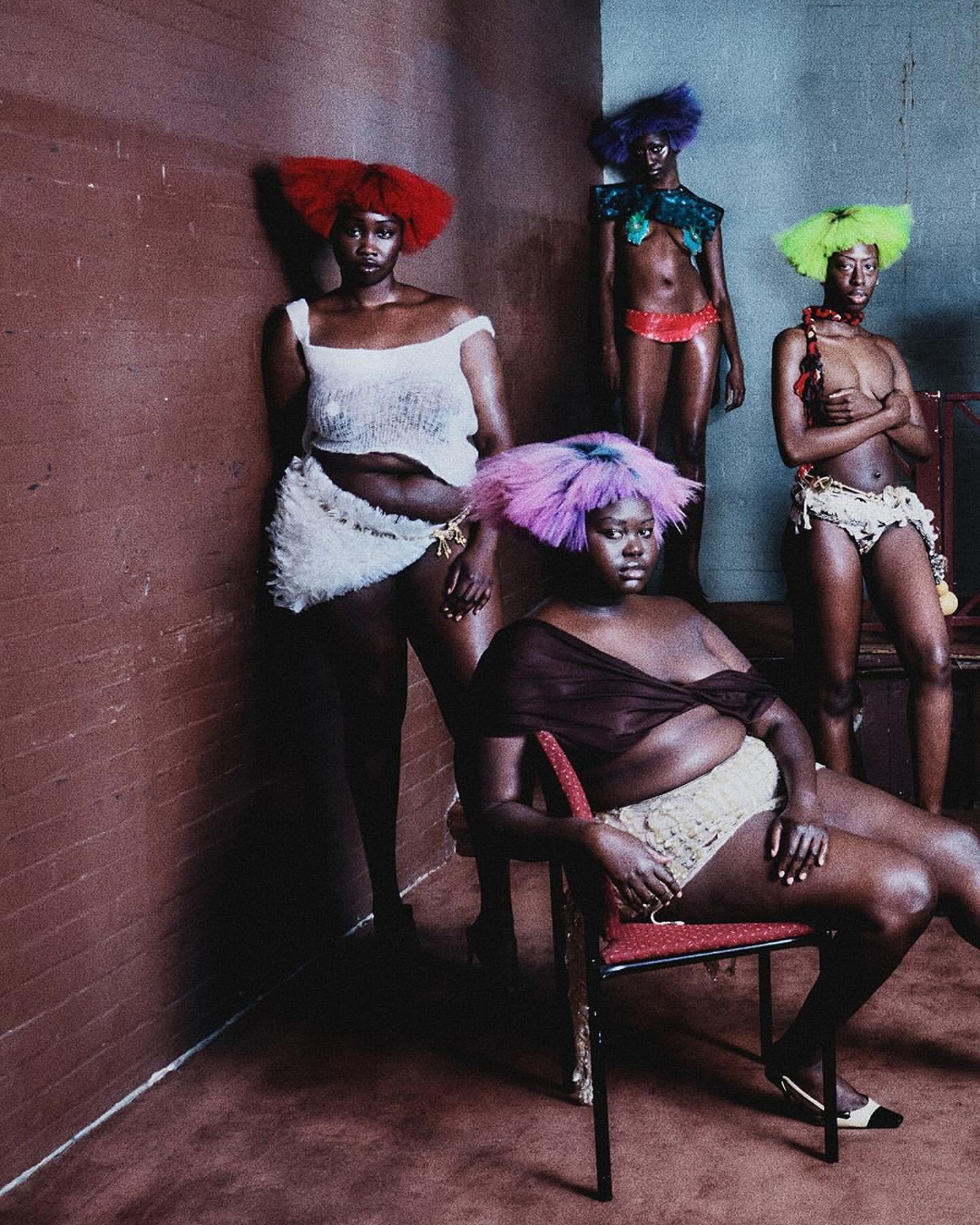 Group of five dark-skinned people in colorful wigs and unique clothing posing in a room with reddish-brown walls, creative fashion editorial.
