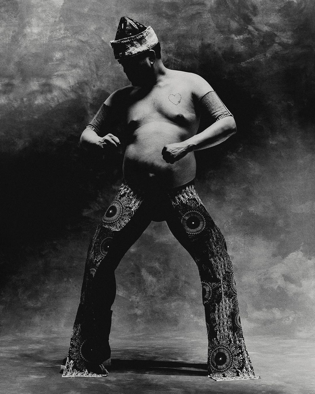 Monochrome studio portrait of a single adult male in embellished pants and headdress, striking a dynamic pose against a mottled backdrop.