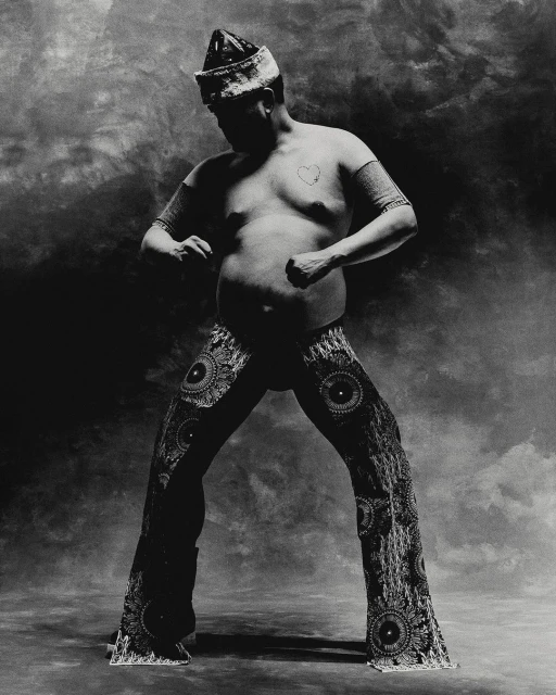 Monochrome studio portrait of a single adult male in embellished pants and headdress, striking a dynamic pose against a mottled backdrop.