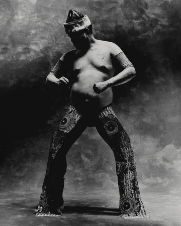 Monochrome studio portrait of a single adult male in embellished pants and headdress, striking a dynamic pose against a mottled backdrop.