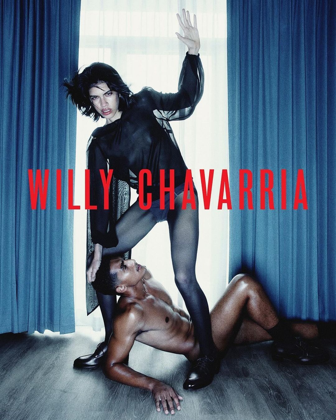 Fashion shot: a standing female model with dark hair places a foot on a shirtless male model in a photo studio setting with text overlay.