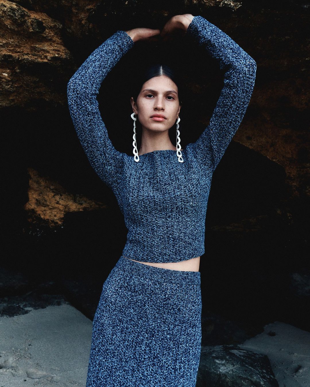 A woman in a knitted top and skirt poses with her hands above her head in an outdoor location in front of a dark rocky backdrop.