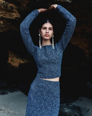 A woman in a knitted top and skirt poses with her hands above her head in an outdoor location in front of a dark rocky backdrop.