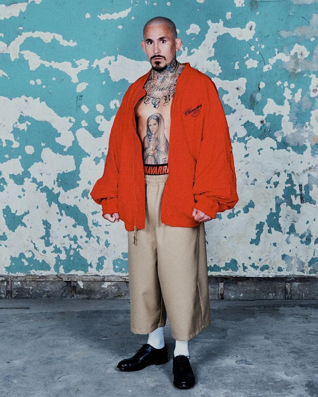 Man with tattoos wears a red jacket, beige pants, and black dress shoes against a distressed blue-and-white wall, showcasing an edgy style.
