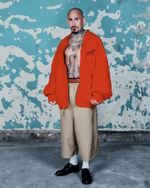Man with tattoos wears a red jacket, beige pants, and black dress shoes against a distressed blue-and-white wall, showcasing an edgy style.