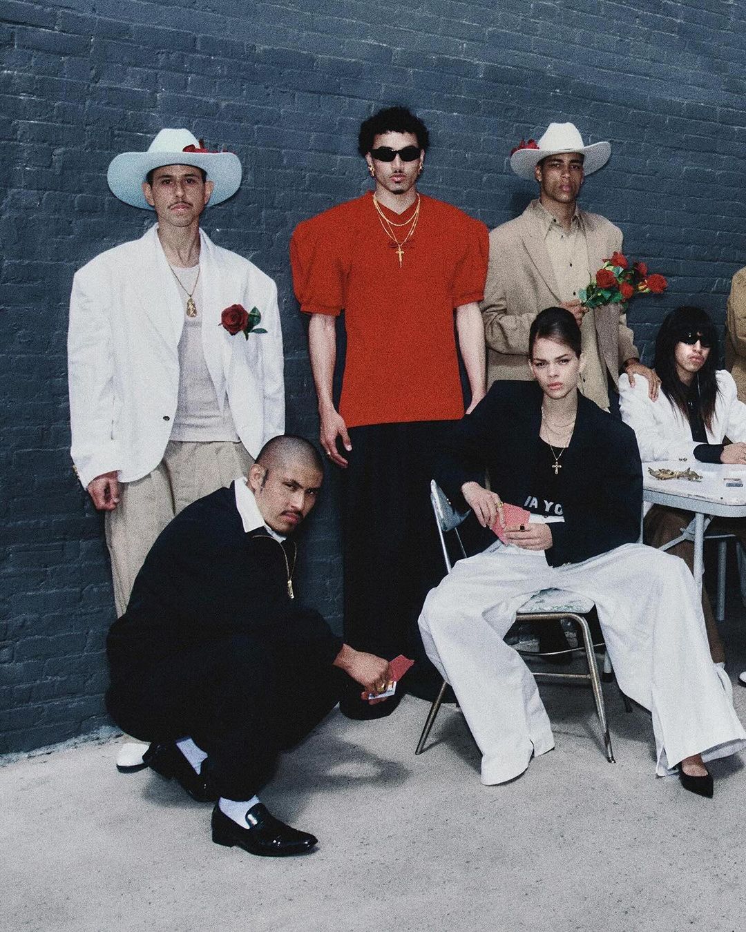 Group of people in an indoor setting with black brick wall backdrop, diverse clothing styles, including hats and jewelry, and some holding playing cards.