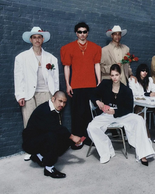 Group of people in an indoor setting with black brick wall backdrop, diverse clothing styles, including hats and jewelry, and some holding playing cards.