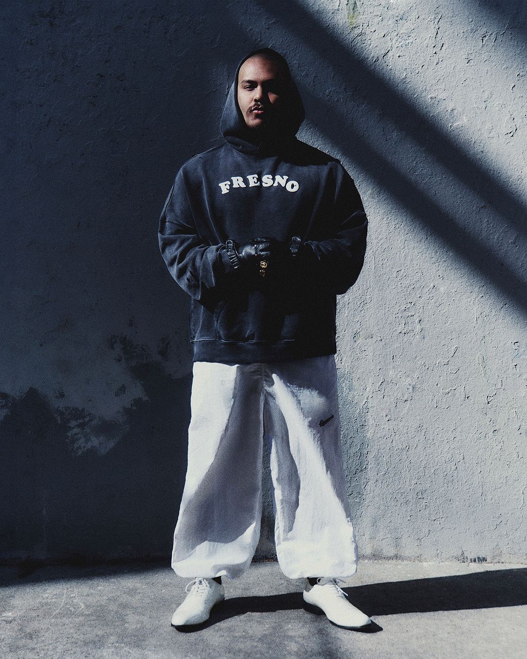Man wearing a dark Fresno hoodie, white trousers, and white shoes standing against a textured concrete wall with angled shadows.