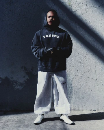 Man wearing a dark Fresno hoodie, white trousers, and white shoes standing against a textured concrete wall with angled shadows.