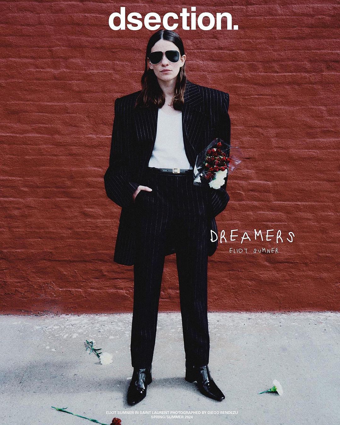 An adult fashion model poses standing against a brick wall, wearing a black pinstripe suit and sunglasses while holding a bouquet of red flowers.