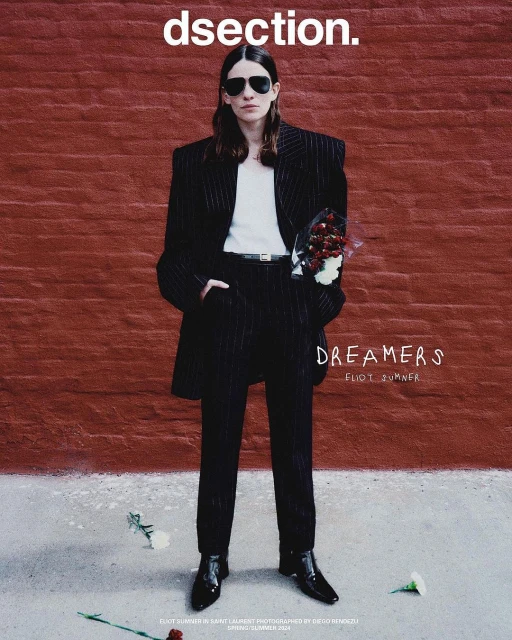 An adult fashion model poses standing against a brick wall, wearing a black pinstripe suit and sunglasses while holding a bouquet of red flowers.
