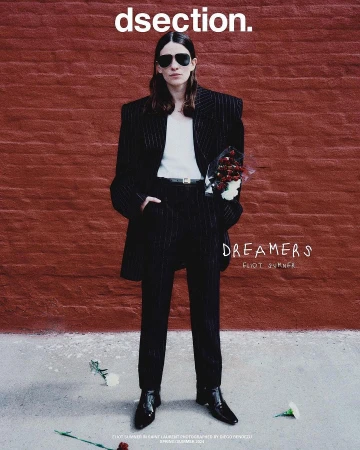 An adult fashion model poses standing against a brick wall, wearing a black pinstripe suit and sunglasses while holding a bouquet of red flowers.