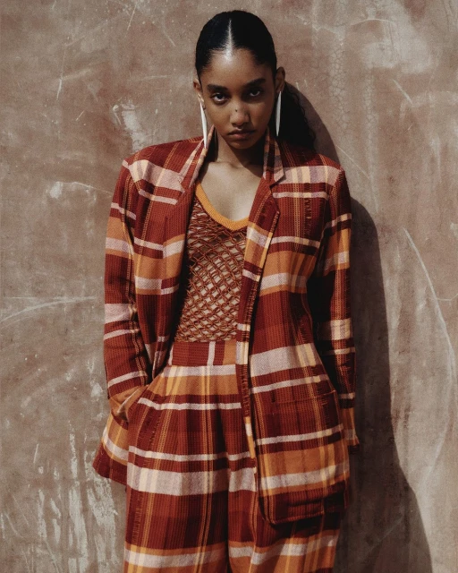 Woman in plaid suit poses indoors against textured wall in portrait. She wears earrings and a sheer top with a V-neck.