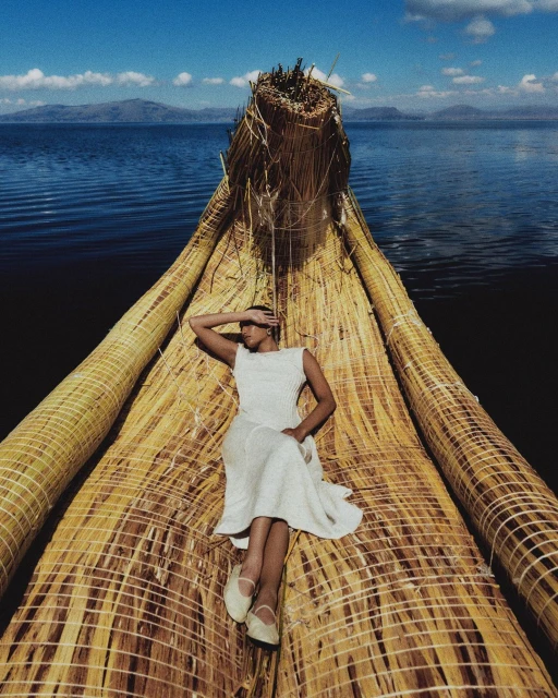 Woman in white dress lying on a straw boat floating on a blue lake against a mountain backdrop, creating a serene travel image.