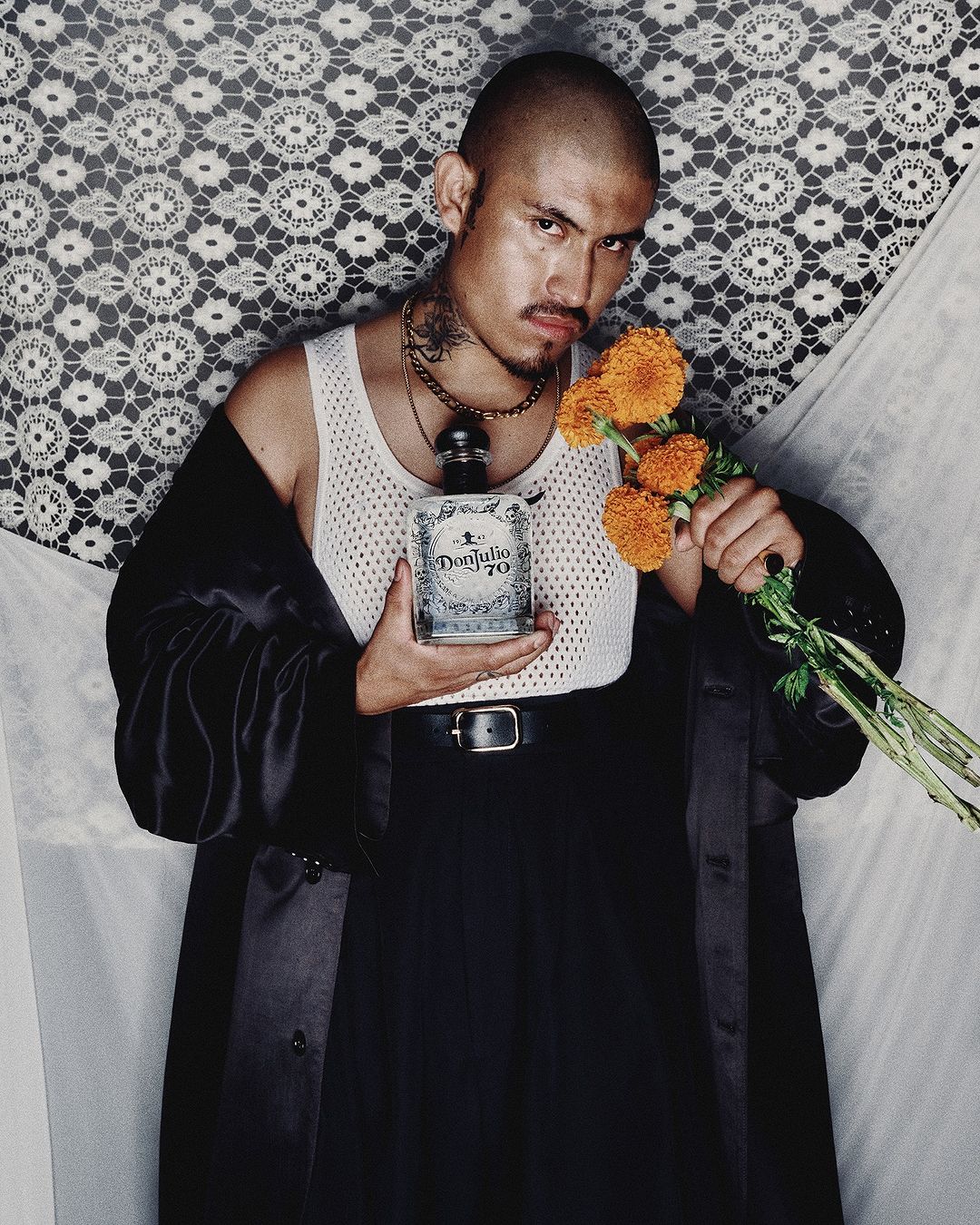 Adult male posing with a bottle of tequila and orange flowers. He is wearing a white undershirt and a black coat, against a neutral backdrop.