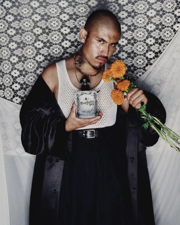 Adult male posing with a bottle of tequila and orange flowers. He is wearing a white undershirt and a black coat, against a neutral backdrop.