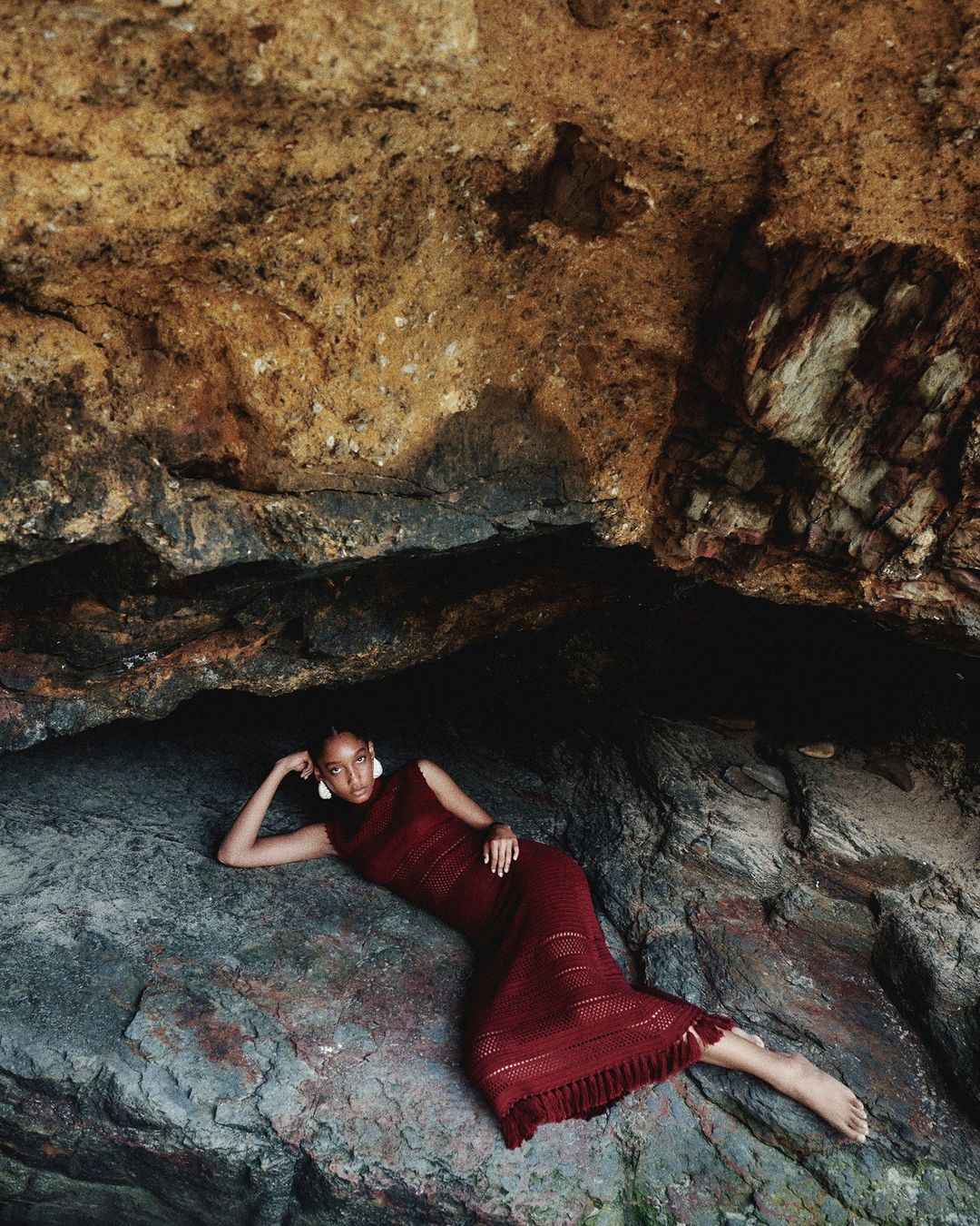 Adult woman in a maroon dress laying on a rock formation inside a cave with neutral tones, wearing white earrings, shot with natural lighting.