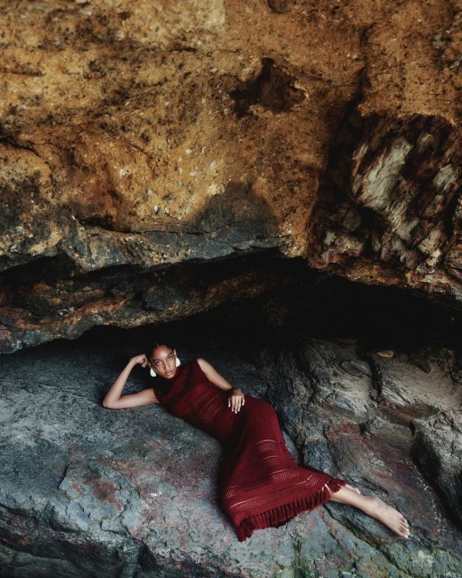 Adult woman in a maroon dress laying on a rock formation inside a cave with neutral tones, wearing white earrings, shot with natural lighting.