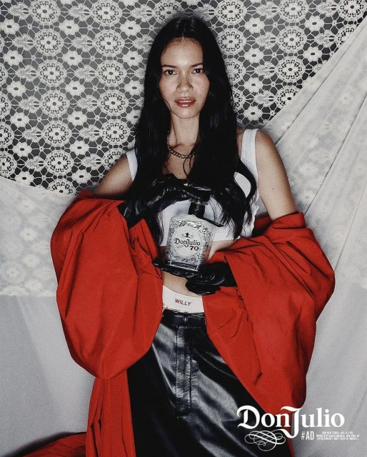 Woman posing with Don Julio 70 tequila bottle in a studio setting with lace backdrop, red jacket, and black leather pants.