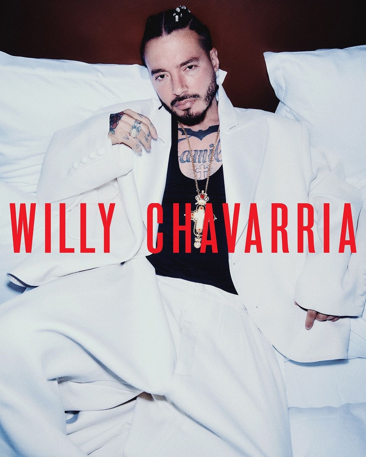 Latin male artist J Balvin poses in a white suit jacket and black top against a white pillow. Willy Chavarria text overlay.