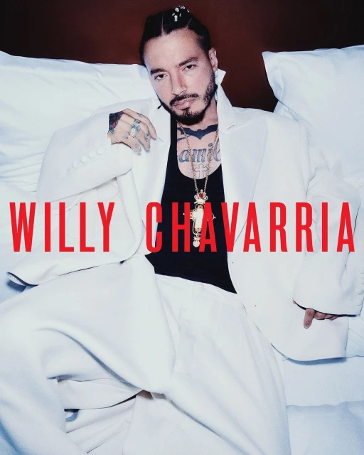 Latin male artist J Balvin poses in a white suit jacket and black top against a white pillow. Willy Chavarria text overlay.