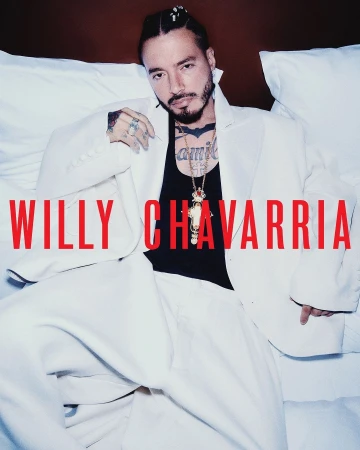 Latin male artist J Balvin poses in a white suit jacket and black top against a white pillow. Willy Chavarria text overlay.