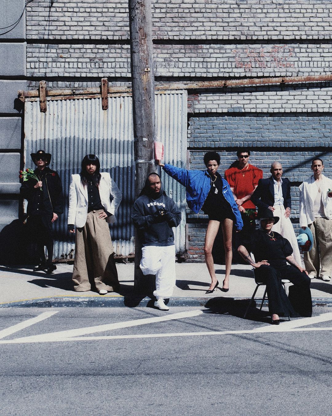 Group of eight people, in varying attire, pose on a city street next to a weathered brick building with corrugated metal doors.