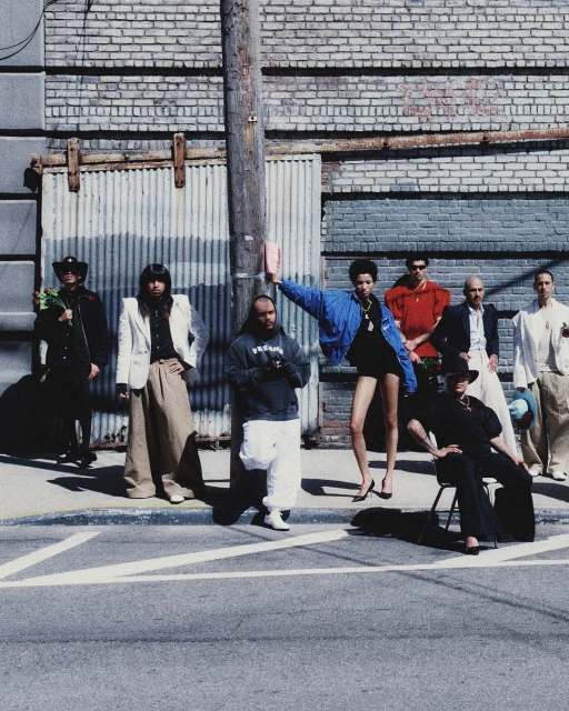 Group of eight people, in varying attire, pose on a city street next to a weathered brick building with corrugated metal doors.