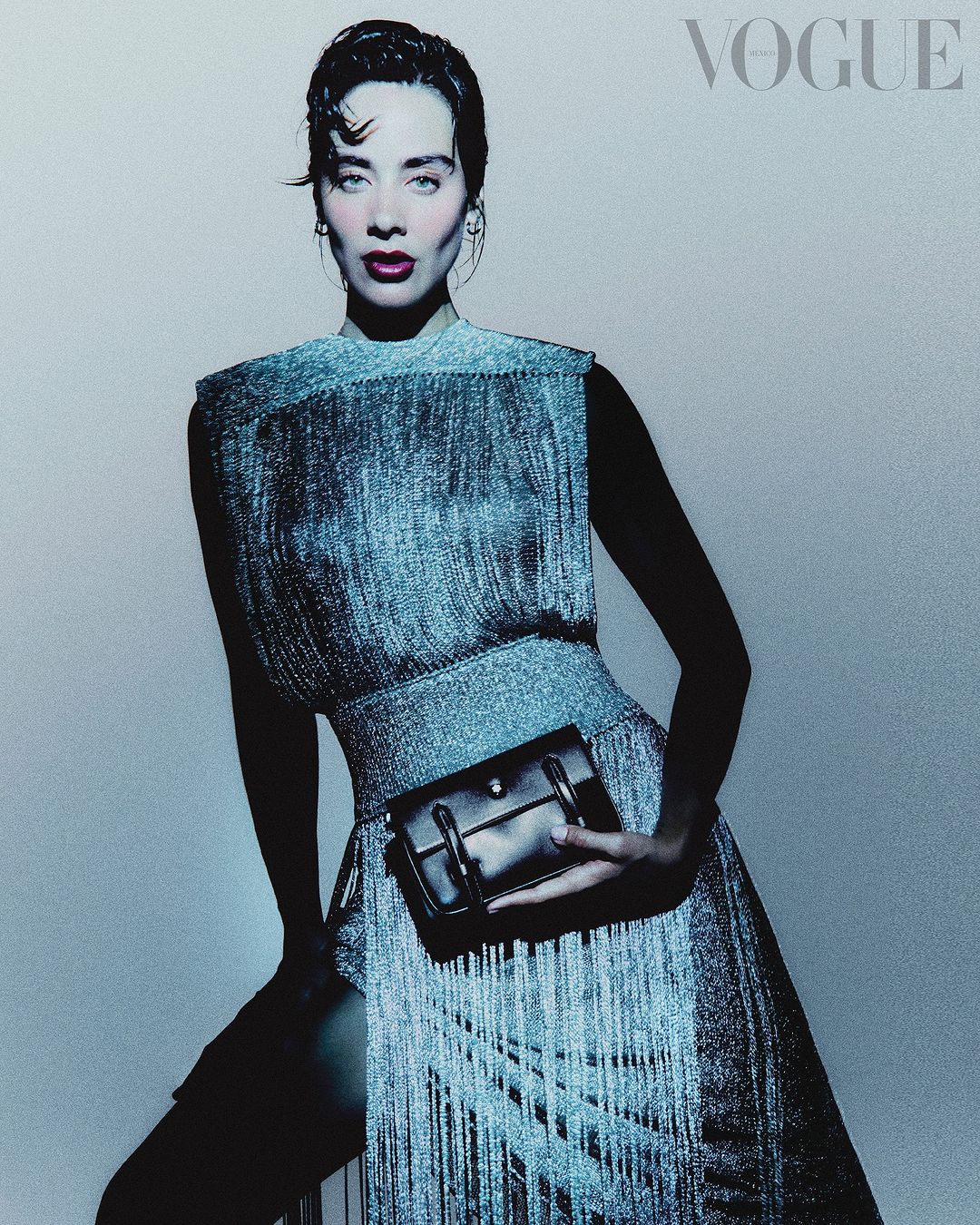 A woman in a shimmering fringe dress poses with a black handbag, against a soft blue backdrop, in a studio setting for a Vogue cover.