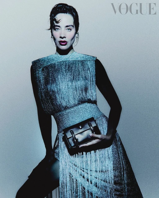 A woman in a shimmering fringe dress poses with a black handbag, against a soft blue backdrop, in a studio setting for a Vogue cover.