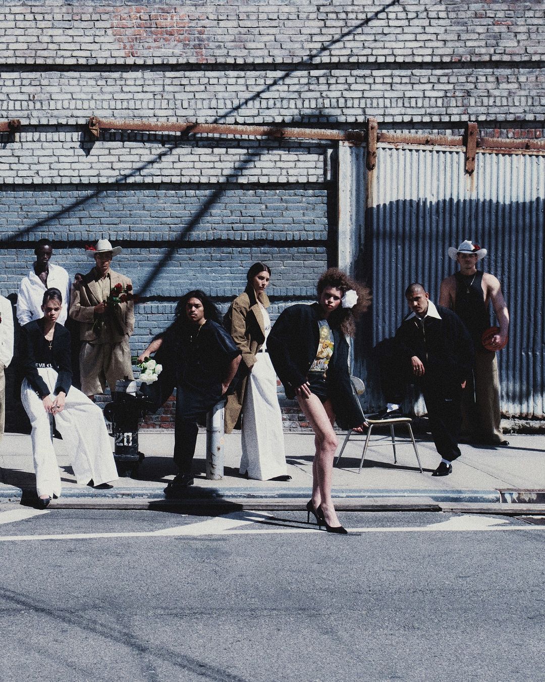Fashion editorial featuring a diverse group of models posing in stylish clothing on a city street with a brick and corrugated metal wall.