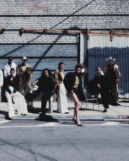 Fashion editorial featuring a diverse group of models posing in stylish clothing on a city street with a brick and corrugated metal wall.