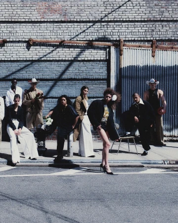 Fashion editorial featuring a diverse group of models posing in stylish clothing on a city street with a brick and corrugated metal wall.