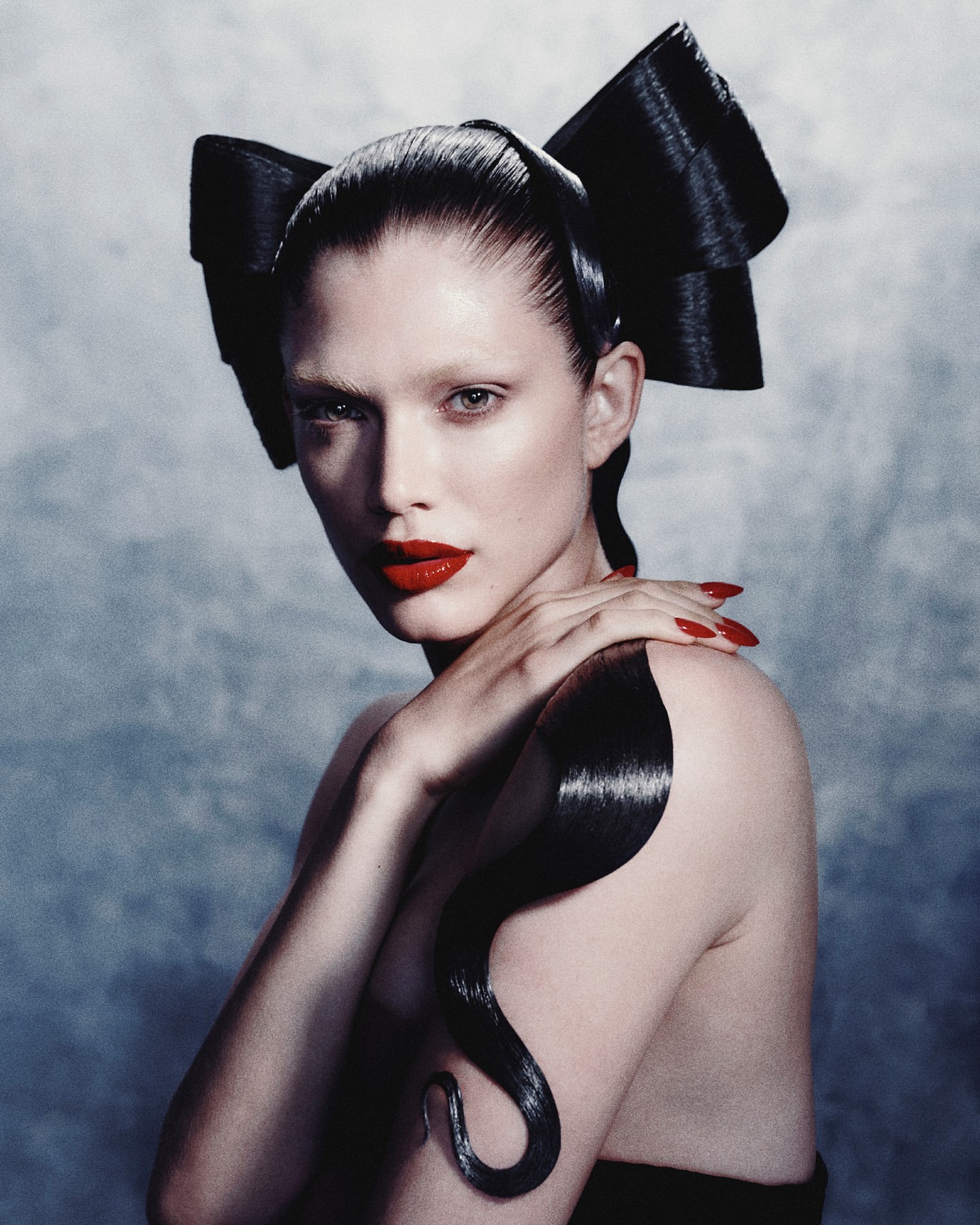 Portrait of a woman with a dramatic black hair bow, bright red lipstick, and shoulder-length curled hairstyle against a grey background.