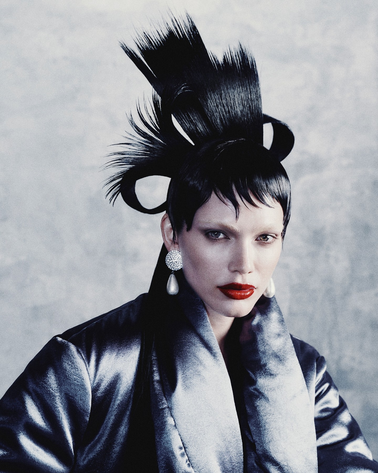 Model with avant-garde black sculpted hair, red lips, and pearl earrings wearing a dark metallic coat against a textured grey background.