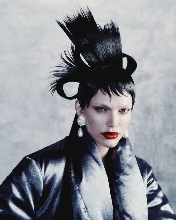 Model with avant-garde black sculpted hair, red lips, and pearl earrings wearing a dark metallic coat against a textured grey background.