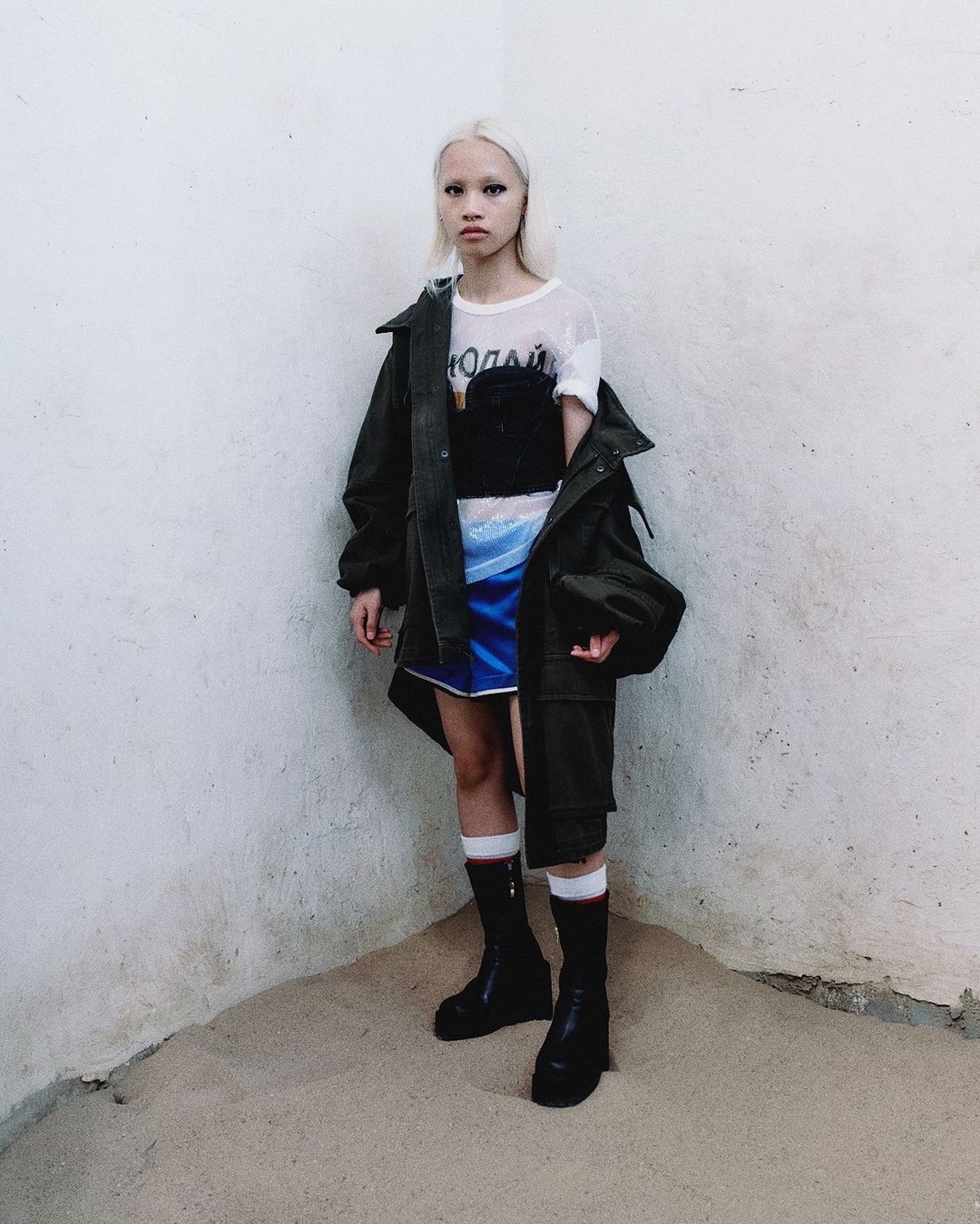 A fair-skinned young adult with platinum blonde hair stands in a corner wearing an olive jacket, a black top, and a blue skirt.