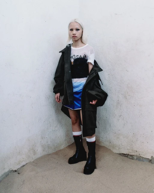 A fair-skinned young adult with platinum blonde hair stands in a corner wearing an olive jacket, a black top, and a blue skirt.