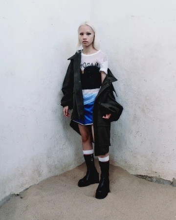 A fair-skinned young adult with platinum blonde hair stands in a corner wearing an olive jacket, a black top, and a blue skirt.