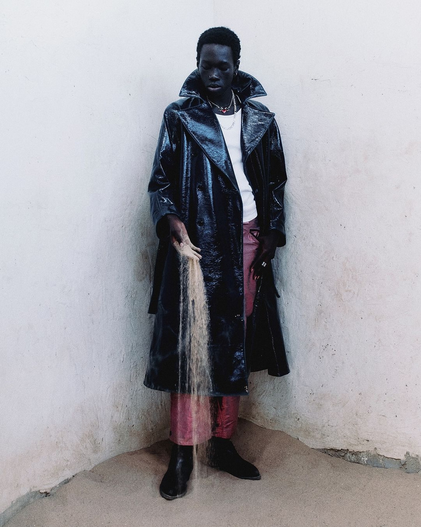 Dark-skinned adult male in a black patent-leather trench coat holds a stream of sand against a white wall in an indoor corner setting.